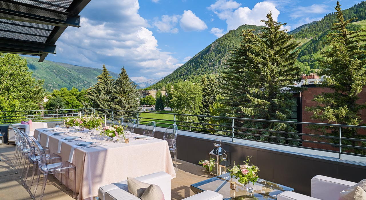 A dining table set for a formal meal on a balcony, surrounded by lush green trees and mountains under a partly cloudy sky, includes elegant floral centerpieces and transparent chairs.