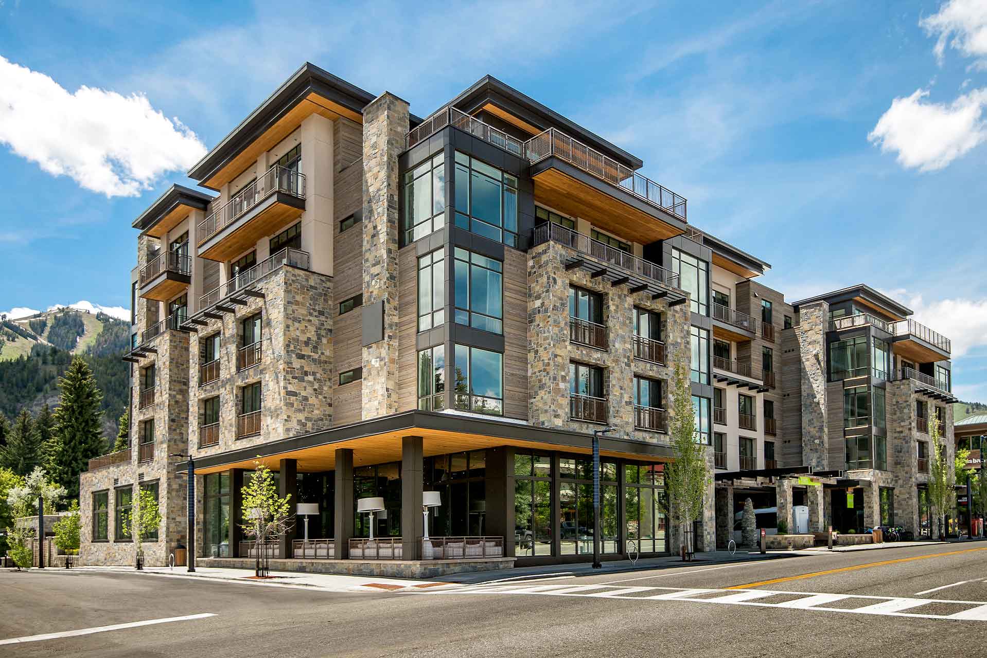 Limelight Ketchum is a modern four-story building with extensive glass windows and stone facade stands on a street corner surrounded by mountains and pine trees under a clear blue sky with a few clouds.