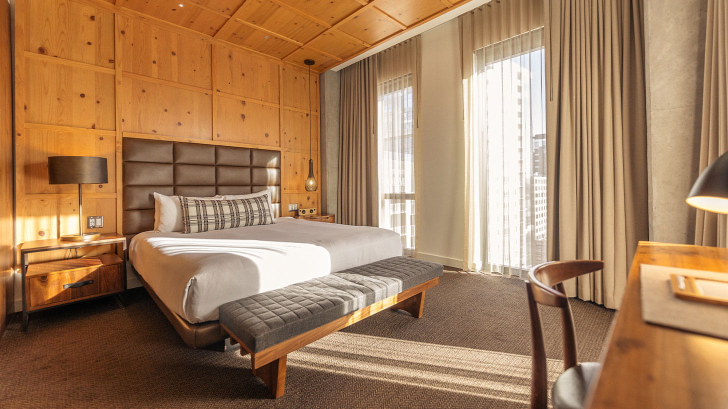 A neatly made bed with a gray headboard and plaid pillows in a sunlit Denver hotel room, with wooden furniture and large windows.