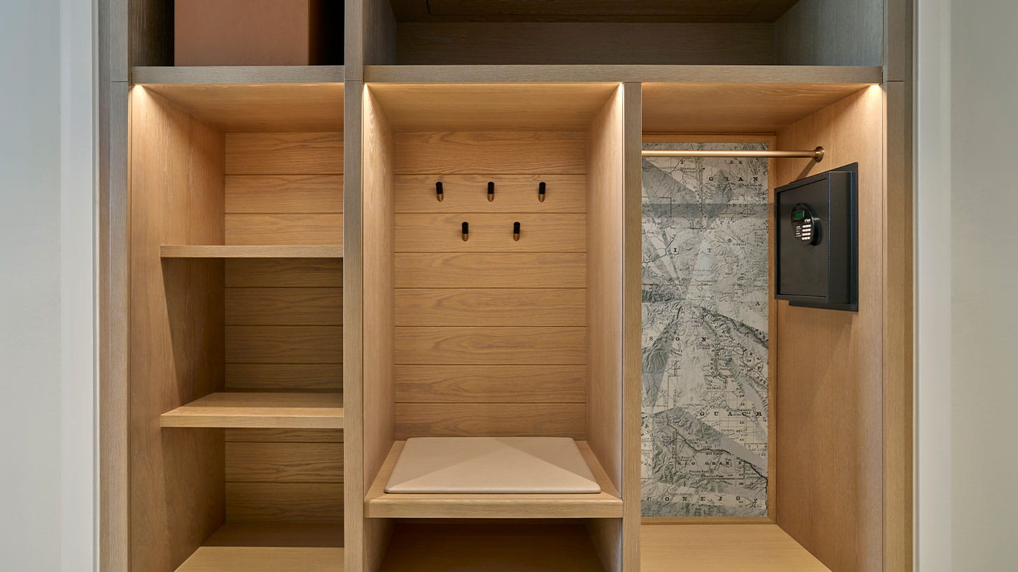 A well-lit wooden ski locker with shelves, hooks, a cushioned bench, a safe, and a hanging bar. A map wallpaper is in the far-right compartment at Aspen.