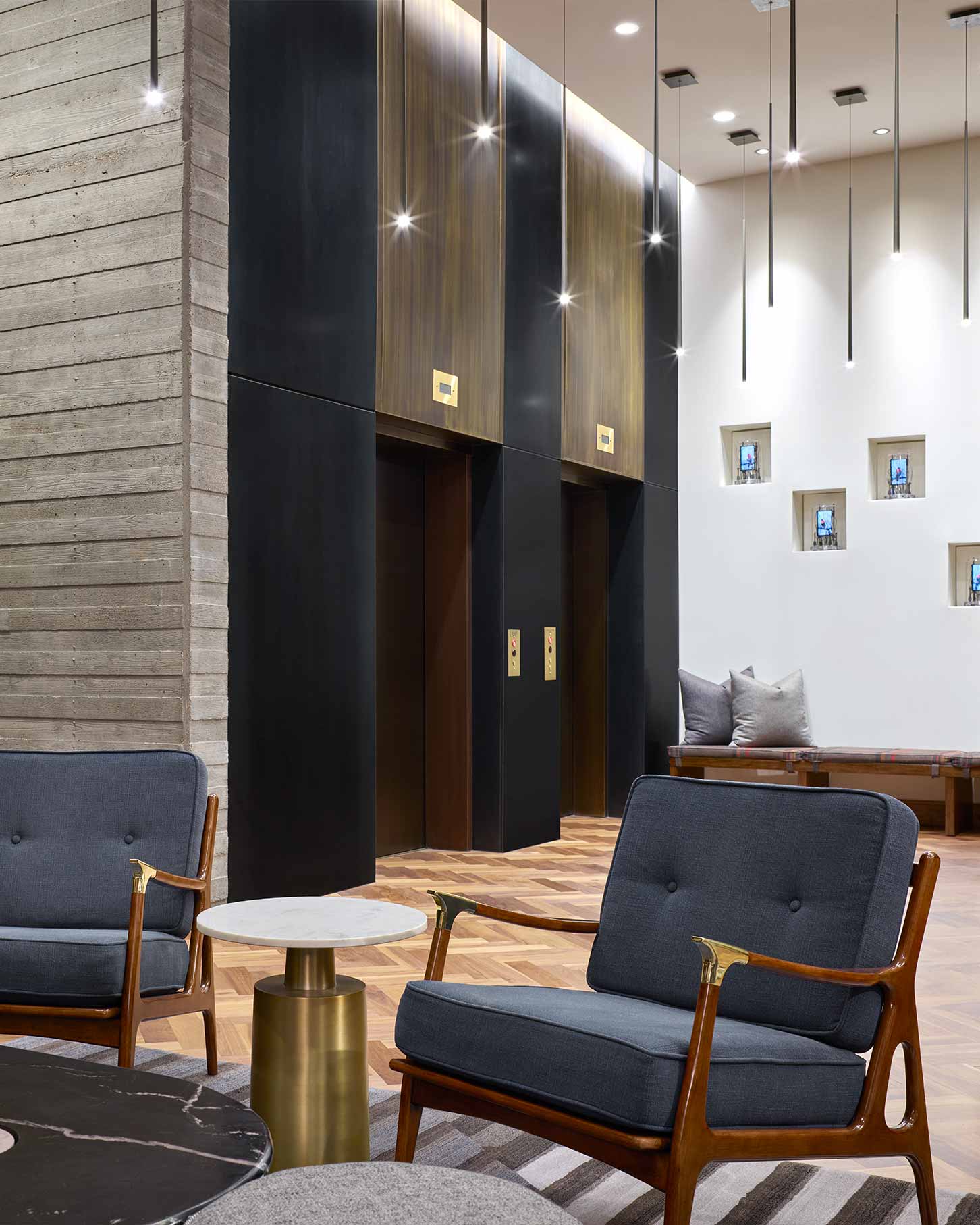 In the Denver lobby, two grey cushioned chairs and a round table sit by a wall with three elevators, floating ceiling lights, and a bench with pillows.