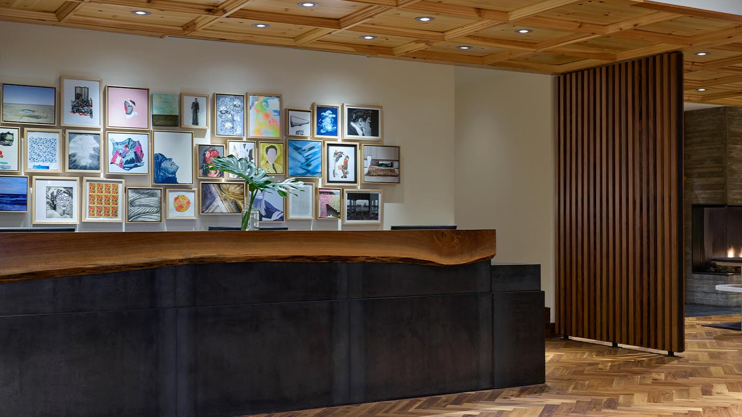 Wooden reception desk at Limelight Denver with a plant in a vase stands before a gallery wall of framed artwork in a modern, warmly-lit room with a wooden ceiling.