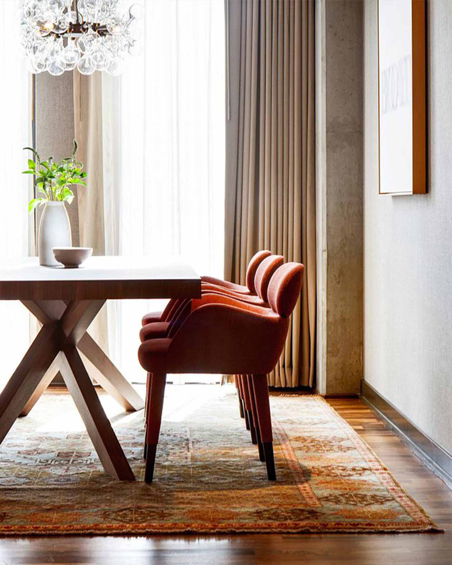 A white dining table with a plant and bowl, surrounded by red chairs, under a chandelier, with curtains and wall art, in Limelight Denver.