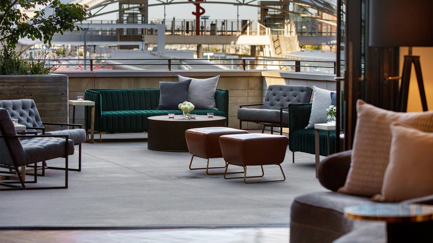 Outdoor lounge in Denver with grey chairs, green sofas, and brown ottomans around a coffee table with flowers and candles, set against modern architecture.