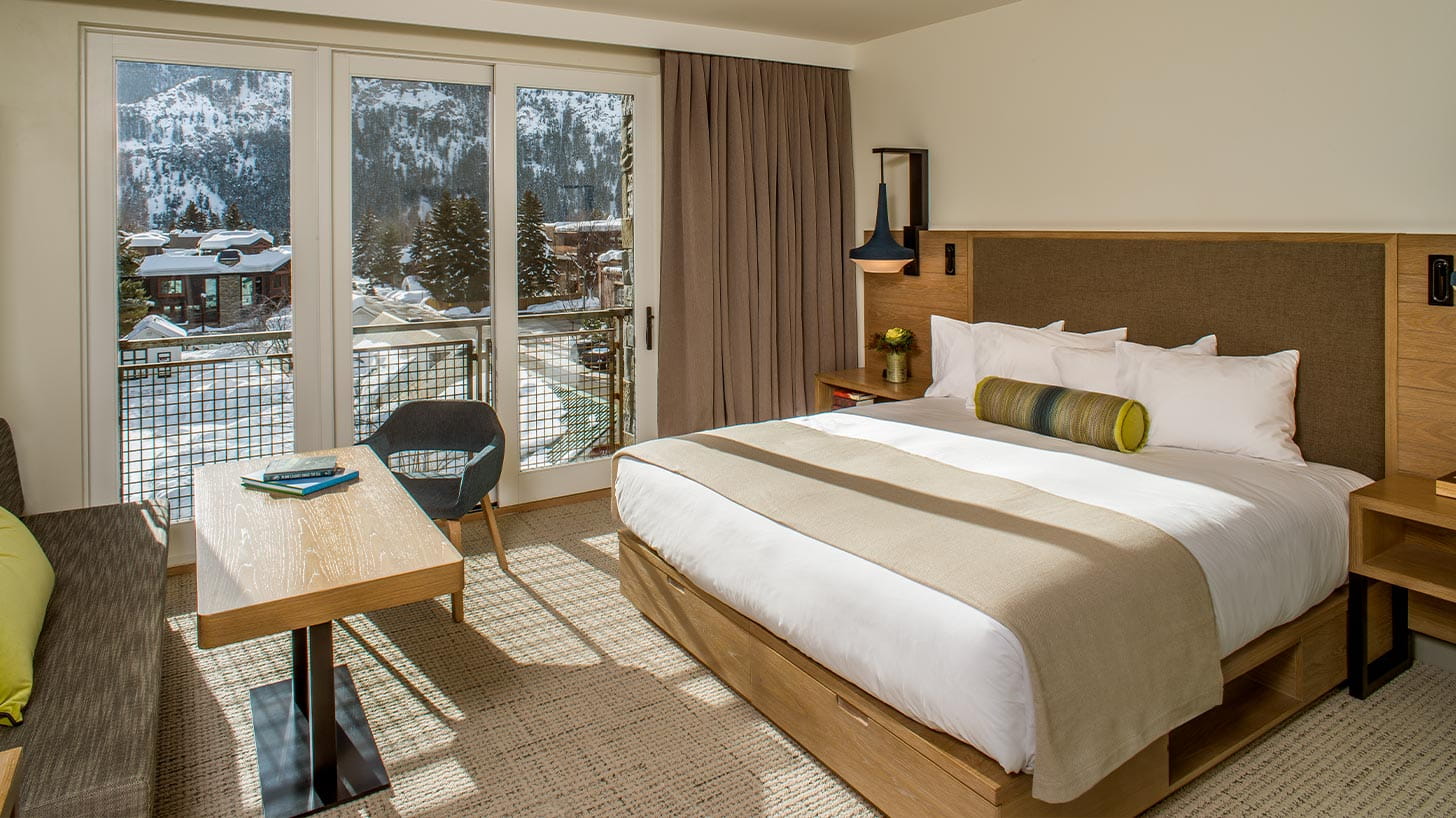 A neatly made bed with white linens in a cozy Limelight Ketchum room, facing a table with books and large windows revealing snowy mountains.