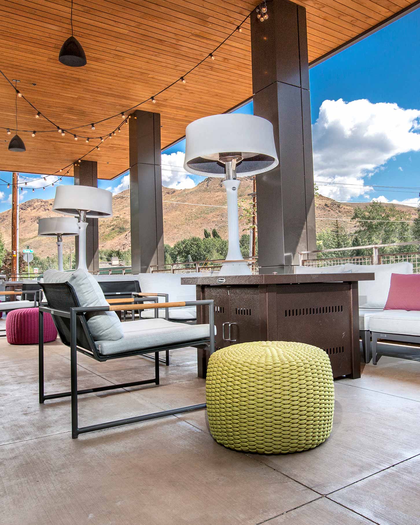 Outdoor lounge in Ketchum with cushioned chairs, colorful stools, patio heaters, and string lights under a wooden ceiling, with mountains and a partly cloudy sky.