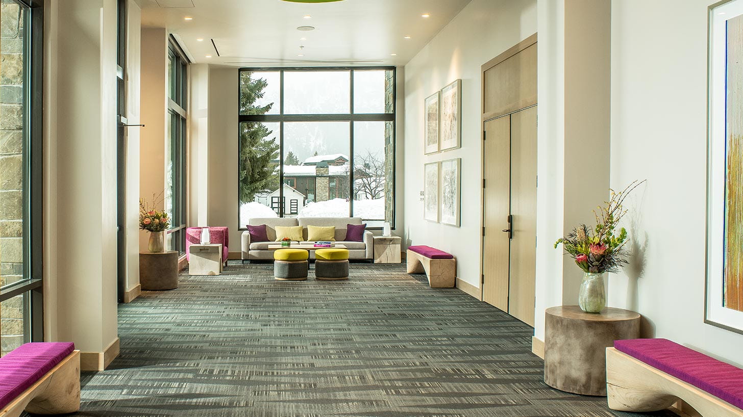 Modern Ketchum hallway with colorful benches and floral arrangements leads to a seating area with sofas, ottomans, large windows with a snowy view, and framed art.