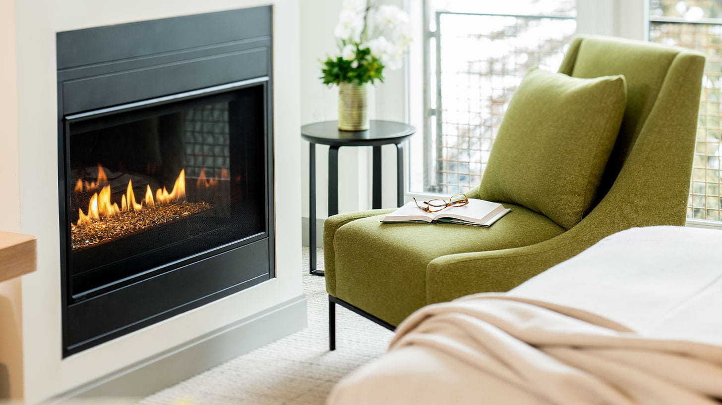 A green chair with an open book and eyeglasses faces a lit fireplace in a cozy living room, with a small side table and potted plant nearby.