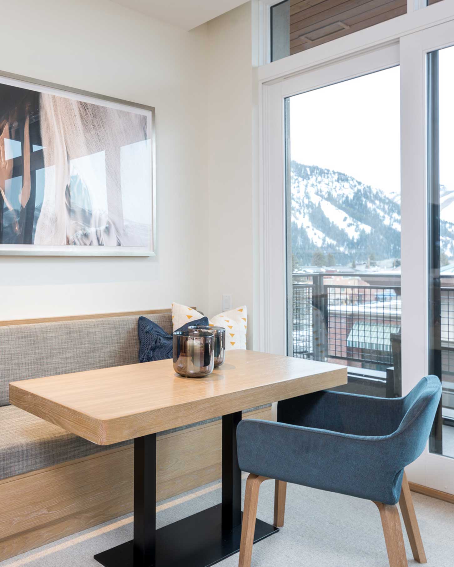 A wooden table with a glass centerpiece sits between a cushioned bench and a blue chair in a modern room with a large window offering a snowy mountain view.