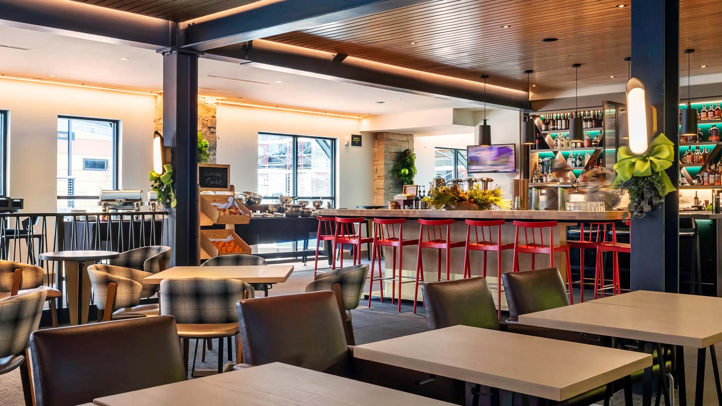 Chairs and tables in a modern Snowmass café with a bar, red stools, an orange fruit stand, and buffet stations, enhanced by bright lighting and decor.