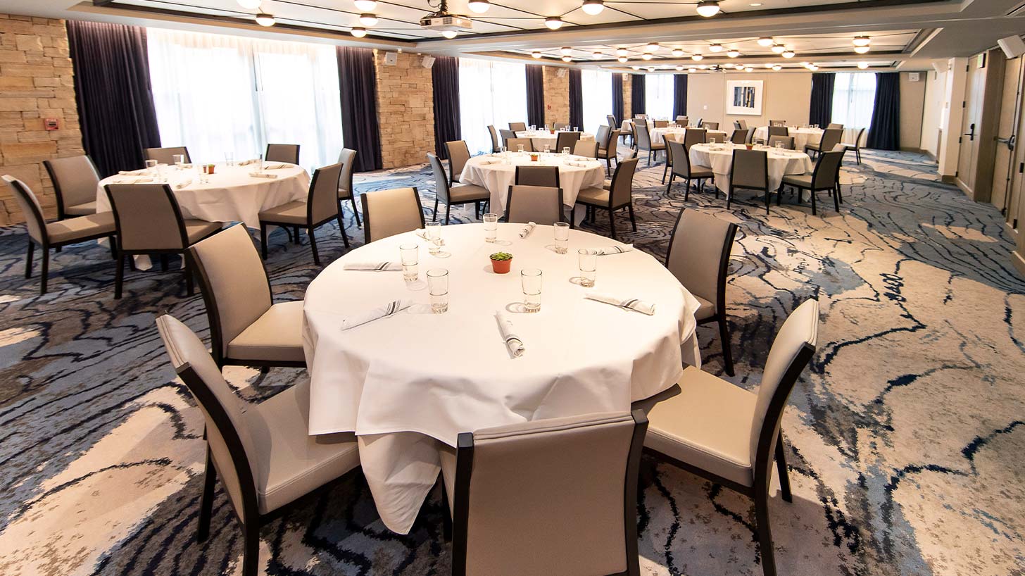 Round tables in a meeting room with white cloths, glassware, and napkins, surrounded by beige chairs, in a well-lit, carpeted event space with stone walls and curtains.