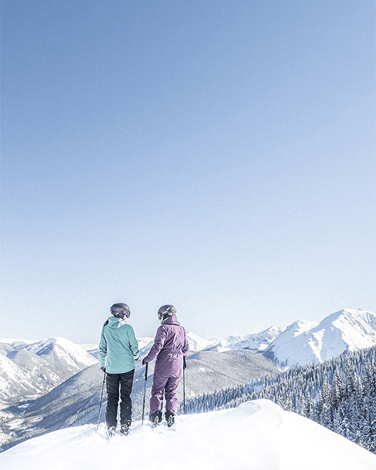 two people stand at the top of a lookout on their skies, soon a beautiful bluebird day as Aspen Snowmass