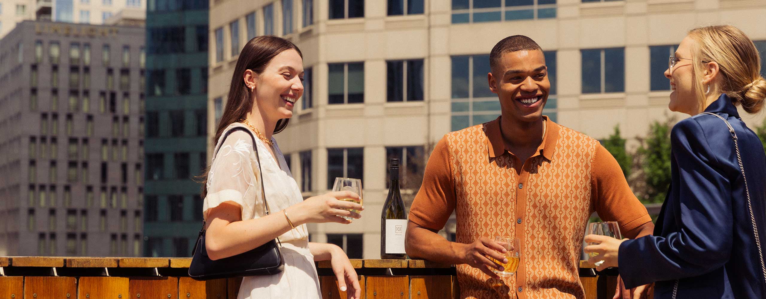 Three people on a Denver rooftop, holding glasses and smiling by a wooden railing with city buildings in the background and a bottle of wine.