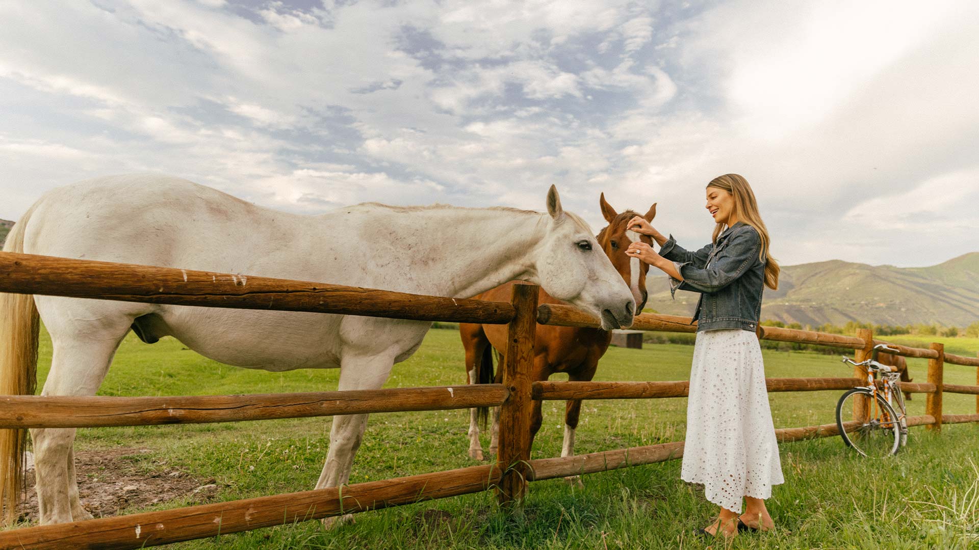 Woman petting two horses in a fenced meadow with mountains in the background.